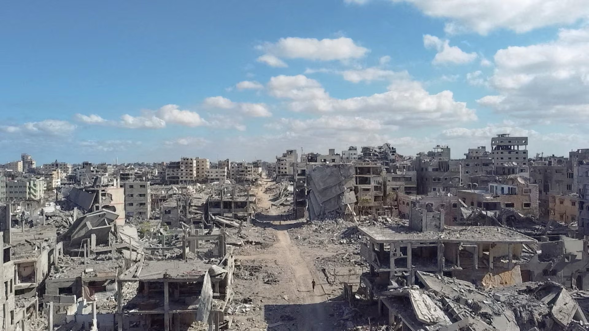 Wide view of Gaza City on October 18 showing heavily damaged and collapsed buildings lining a debris-covered road beneath a blue sky.